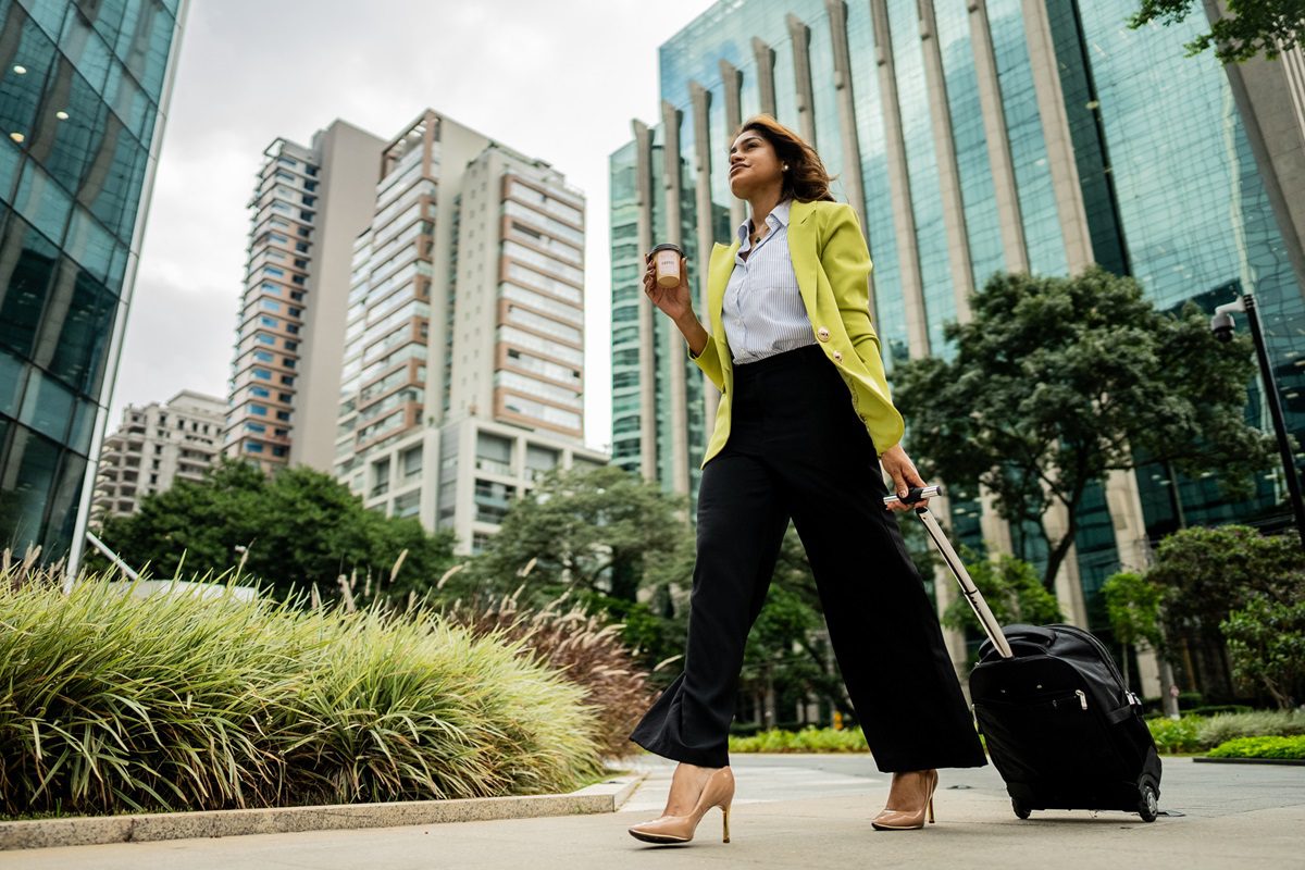 Shot from below, a businesswoman holds a coffee and rolls a suitcase down a city sidewalk