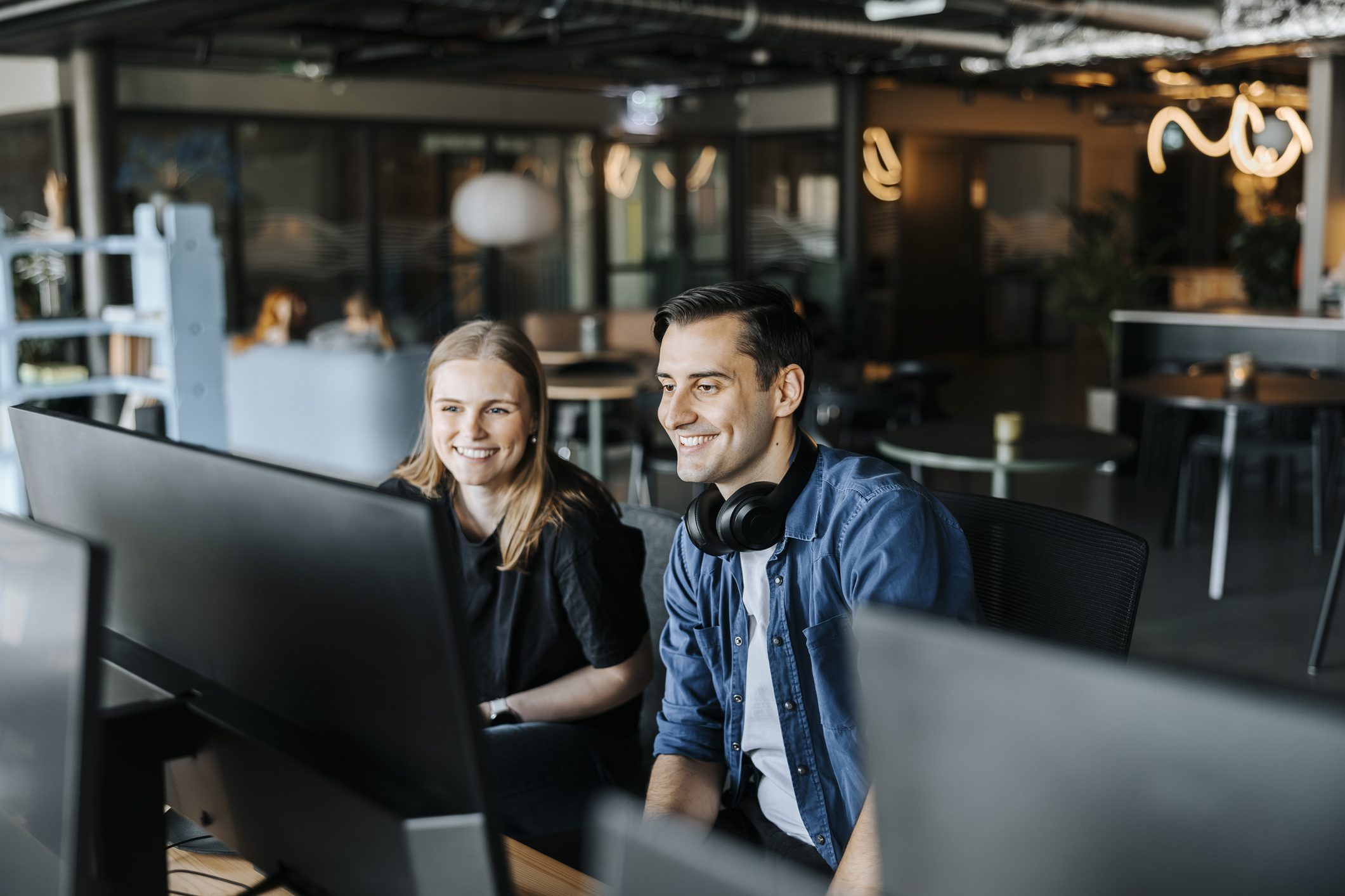 Man and woman sit behind a row of computer monitors in an office, smiling at something on the screen