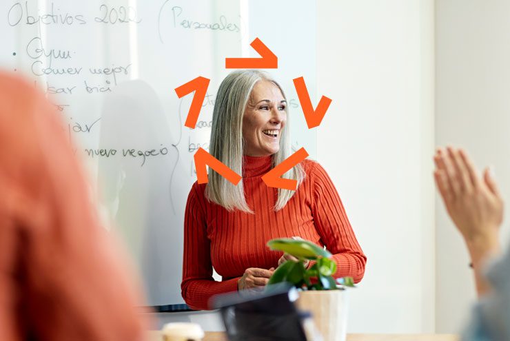 Woman with long hair and an orange top smiles in front of a whiteboard. An Aprio logo has been superimposed around her face.