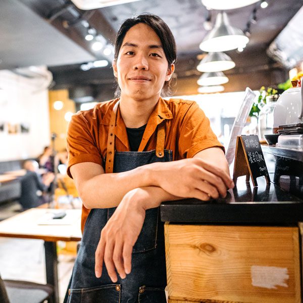 Man with long hair leans against the counter in his cafe and smirks for the camera