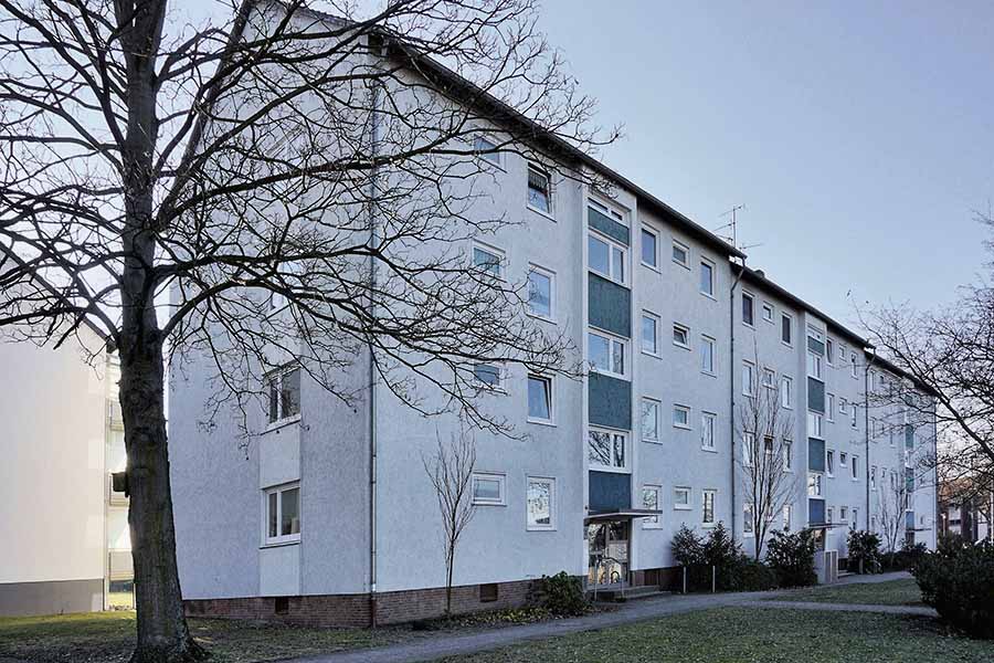 A four-story white apartment building with multiple windows, a sloped roof, and bare trees in front, photographed on a clear day.