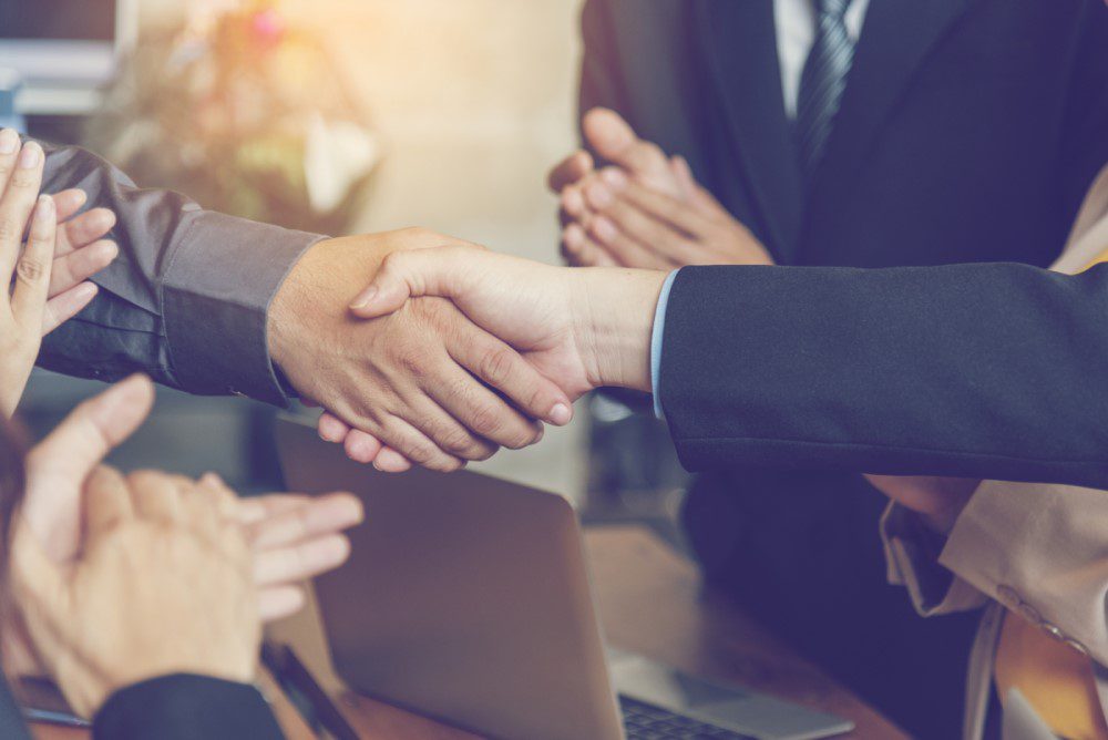 Two people in business attire shake hands across a table, while others nearby clap. A laptop and documents are visible on the table.