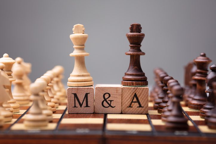 Two chess kings stand on letter blocks reading M & A, surrounded by other chess pieces on a chessboard, symbolizing mergers and acquisitions.