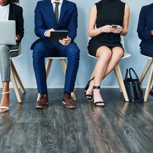 Three people in business attire sit in a row on chairs, using electronic devices. The photo is cropped to show only their bodies from shoulders down.