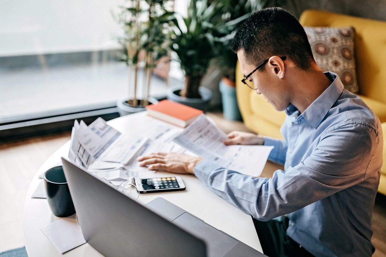 A person in glasses reviews papers at a desk with a laptop, calculator, mug, and plants nearby in a well-lit room.