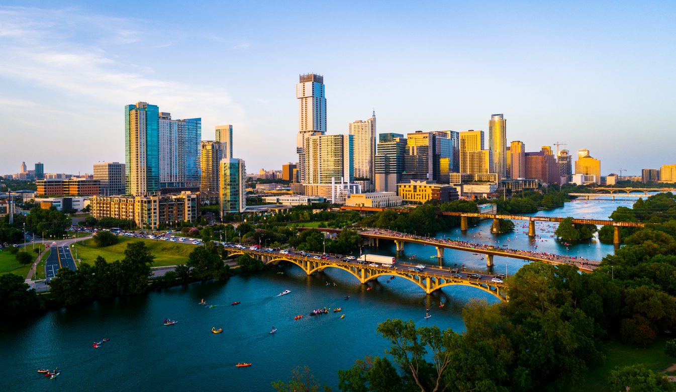 Aerial view of downtown Austin, Texas, showing high-rise buildings, bridges over the river, and people kayaking below on a clear day.
