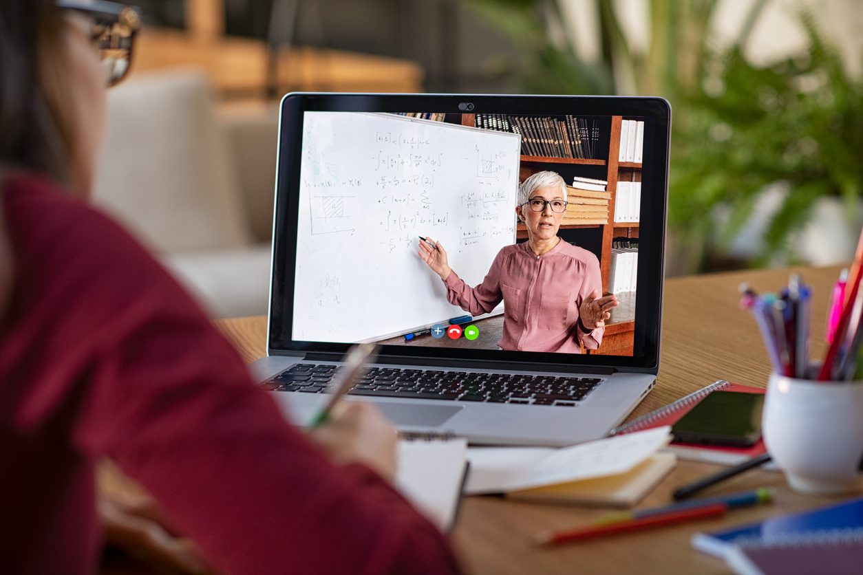 A person takes notes while watching an online lesson; a teacher explains math concepts on a whiteboard during a video call displayed on a laptop screen.