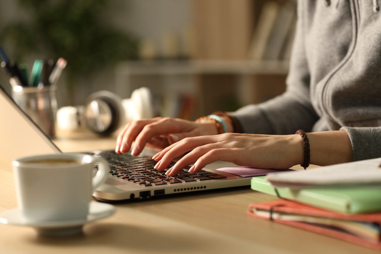 Person typing on a laptop at a desk with a cup of coffee, notebooks, and other office items nearby.
