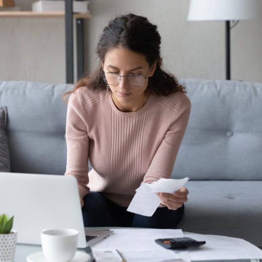 Woman sitting on a couch, looking at a receipt and a laptop, with papers, a calculator, and a cup on the table in front of her.