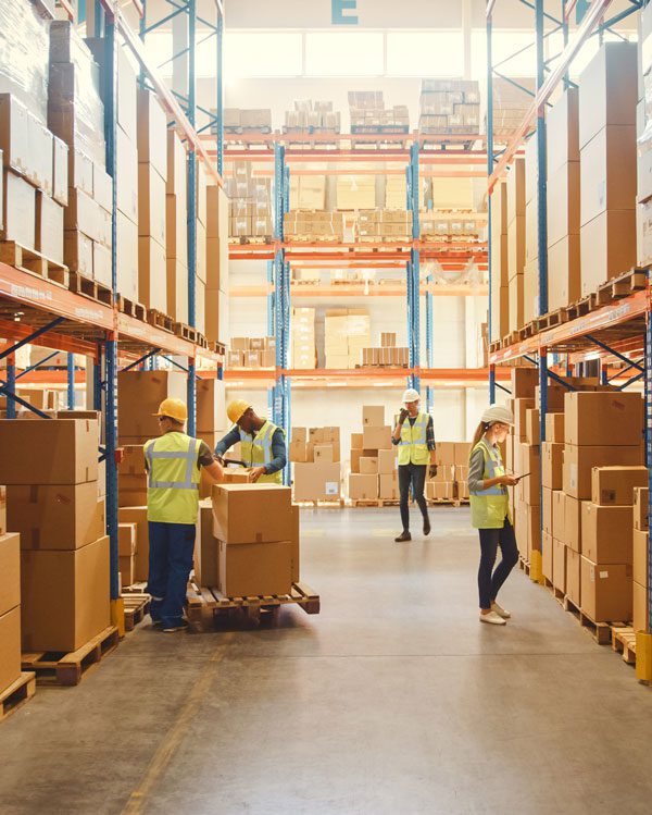 Four workers in reflective vests and hard hats load boxes and take inventory in then aisle of a shipping warehouse