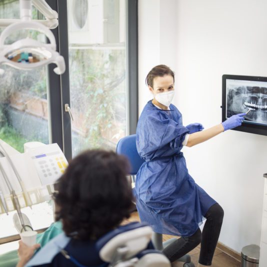 A dentist wearing protective gear points to a dental X-ray on a monitor while explaining it to a patient seated in the dental chair.