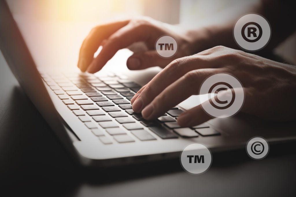 Hands typing on a laptop keyboard with overlaid copyright, trademark, and registered symbols, representing intellectual property concepts.
