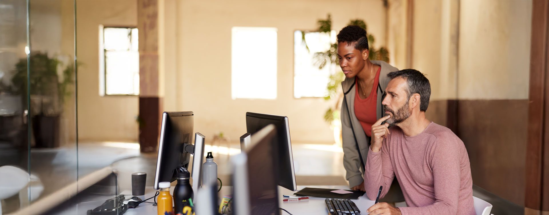 A man sits at a desk and looks at something on a computer while a woman watches from next to him