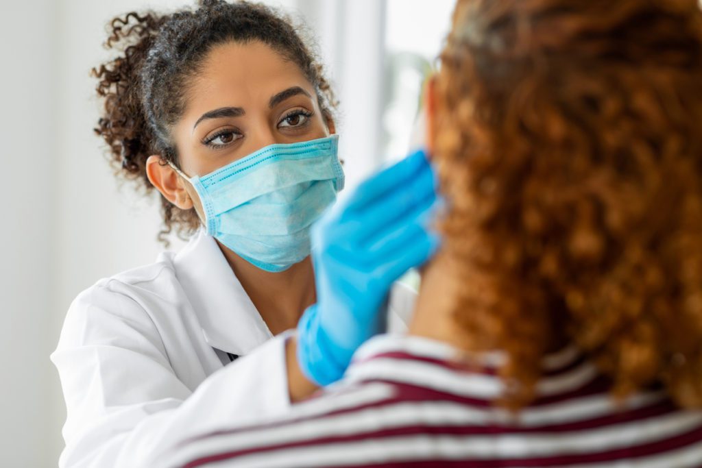 A healthcare professional wearing a mask and gloves examines the face of a patient.