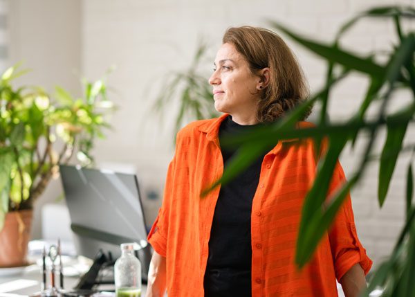 Shot from behind a plant, a woman with short hair and an orange top looks off into the distance