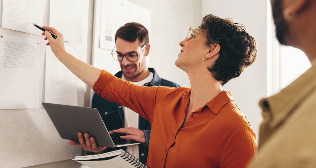 Woman points to a paper on a bulletin board while a man looks at something on a computer