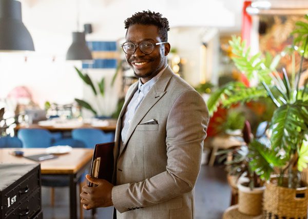 A man in a tan suit jacket stands in a workspace, holds a clipboard under his arm, and smiles at the camera