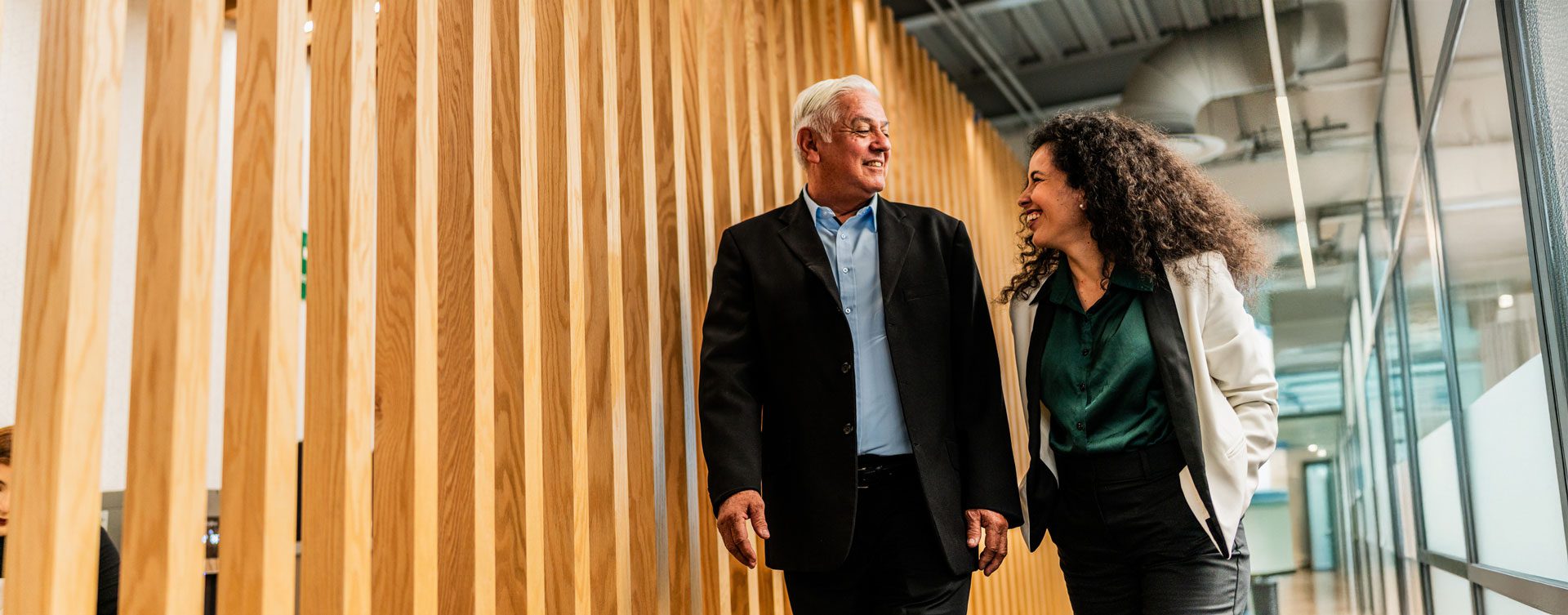 Man and woman laugh and walk down an office hallway lined by wooden slats on one side and glass on the other