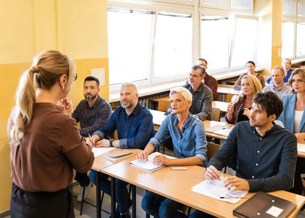 Blonde woman with her back to the camera addresses classroom of men and women who sit at long wooden tables
