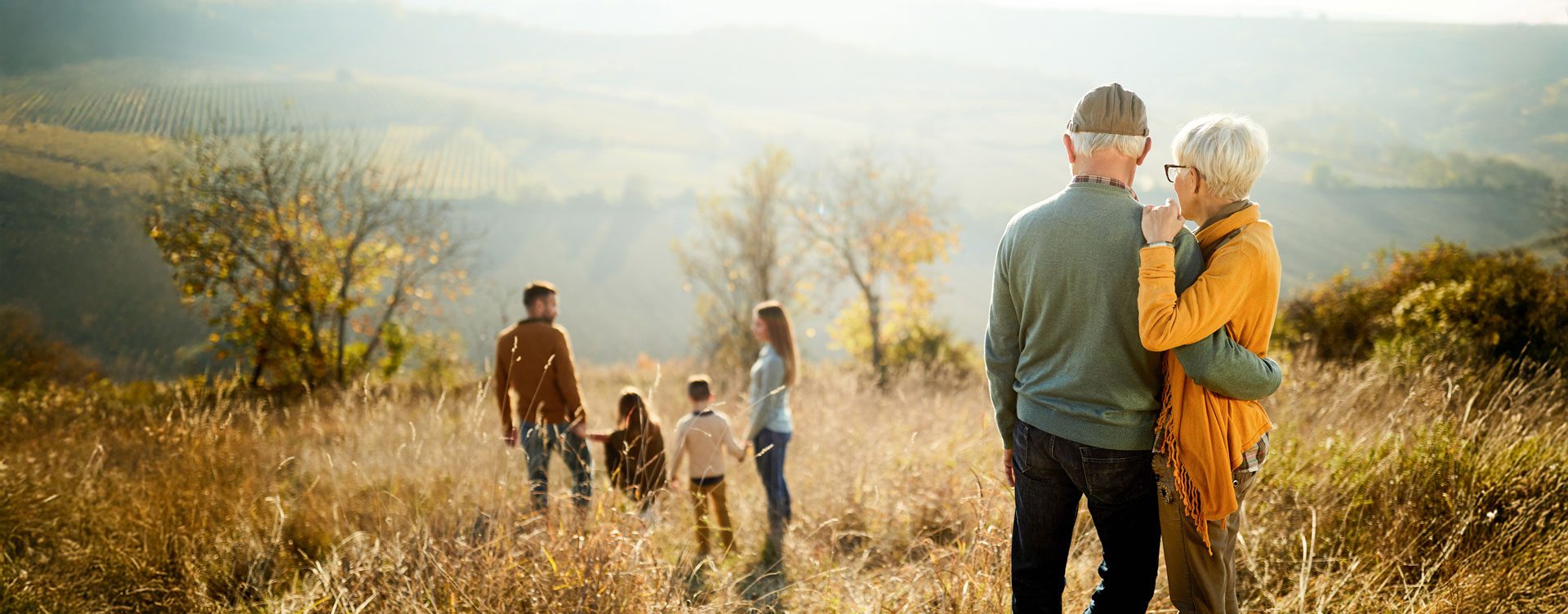 Shot from behind, an old couple watches a young family walk ahead of them through a field on a scenic mountainside