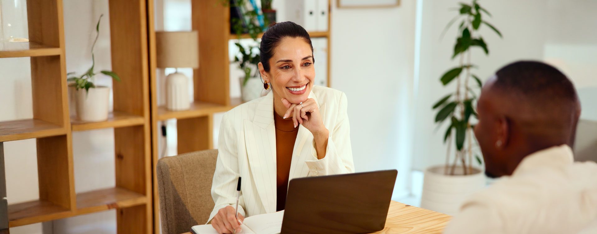 Dark haired woman in a white blazer sits in front of a laptop and smiles at a man blurred in the foreground