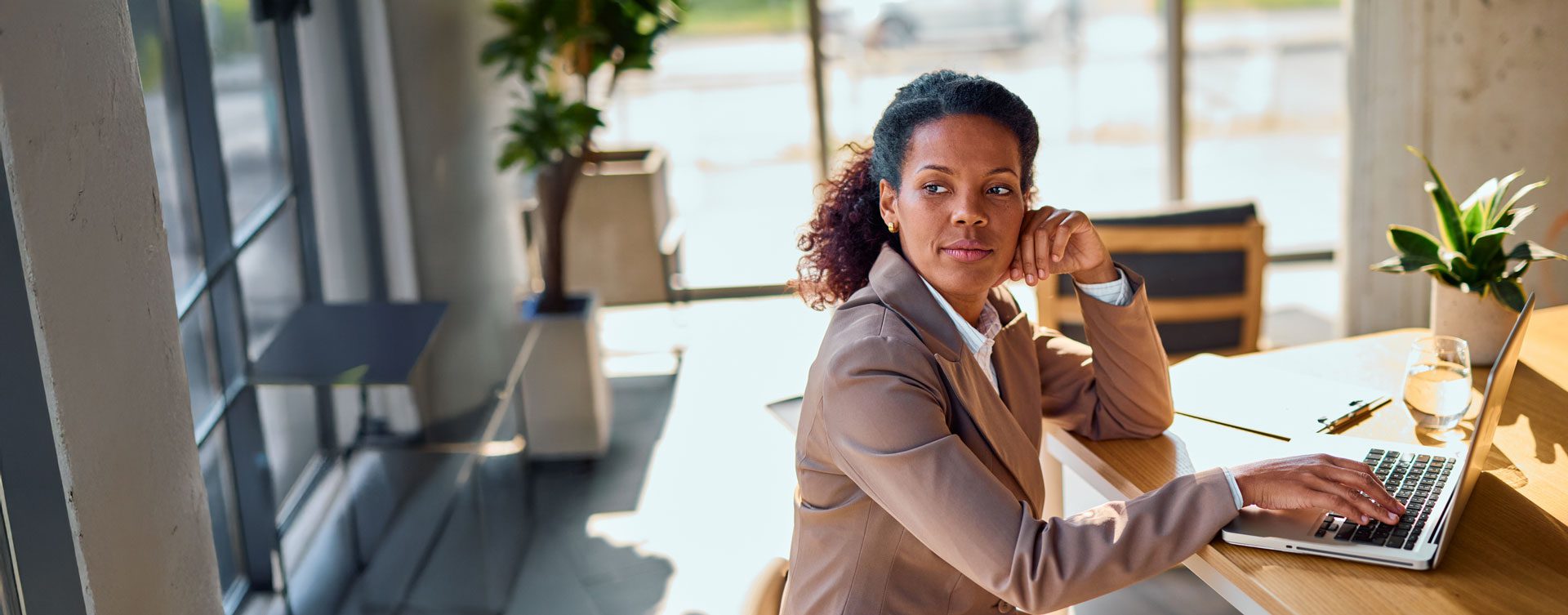 Woman in a brown leather coat uses a laptop at a wooden table and gazes dramatically at something out of frame