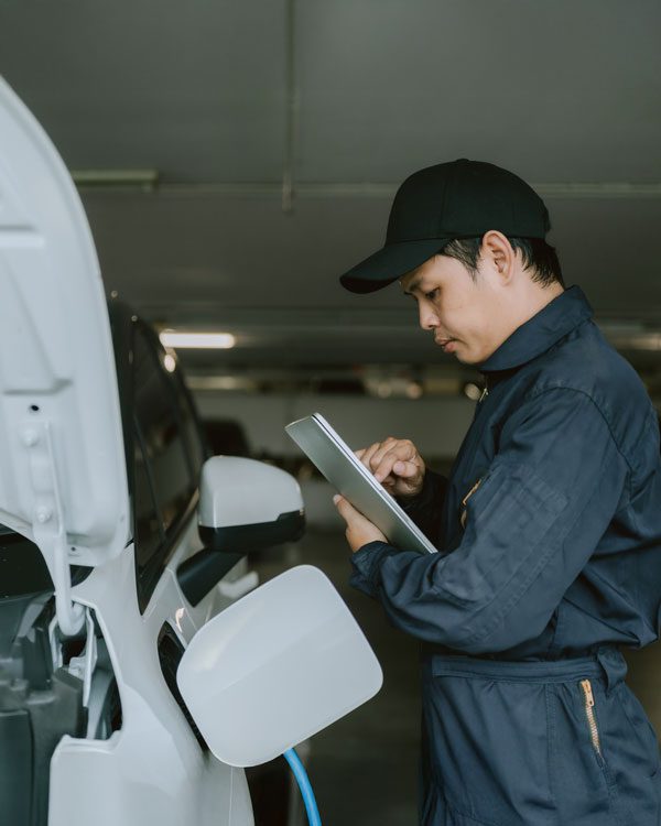 A mechanic in a dark jumpsuit and cap stands beside a white electric vehicle and looks at a tablet