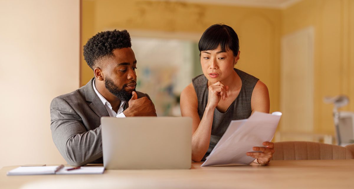 In a yellow room, a man sits at a table in front of a laptop while a woman leans beside him holding papers