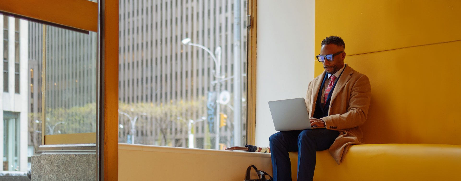 Man in a suit and coat sits on a yellow bench in front of a big window and uses a laptop