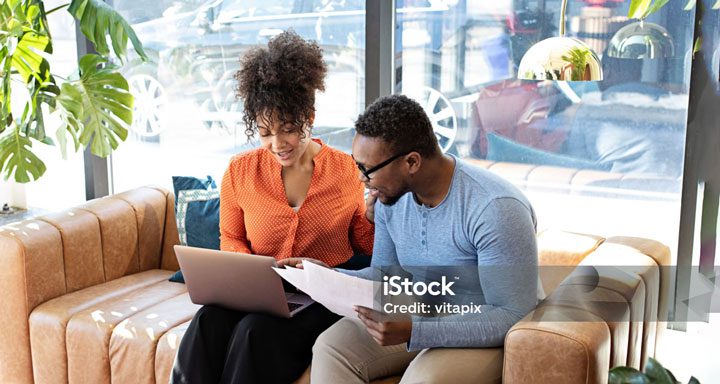 Two people sitting on a sofa, looking at a laptop and reviewing documents together in a bright, modern room.