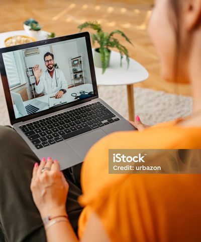 Shot over a woman's shoulder, a doctor sitting in his office waves from a computer screen