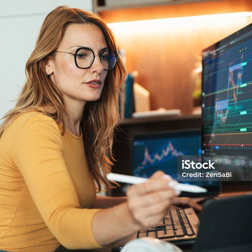 A woman wearing glasses sits at a desk, using a stylus to interact with a computer displaying financial graphs and charts.