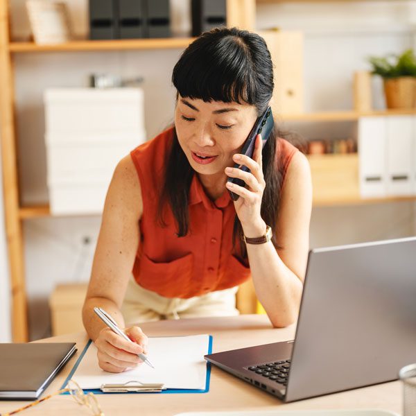 Woman leans against a table and talks on the phone while writing something down on a clipboard
