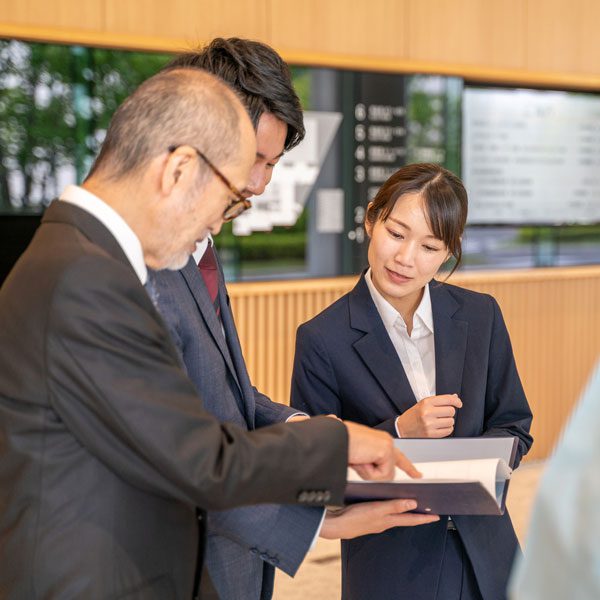 Two businessmen and a businesswoman stand in an office and look over some papers