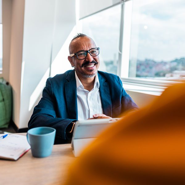 Man in glasses and a suit jacket sits at a desk by a window and smiles at someone obscured in the foreground