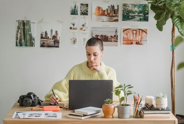 Woman in a yellow sweater sits at a desk in front of a wall of printed out photos and uses a laptio