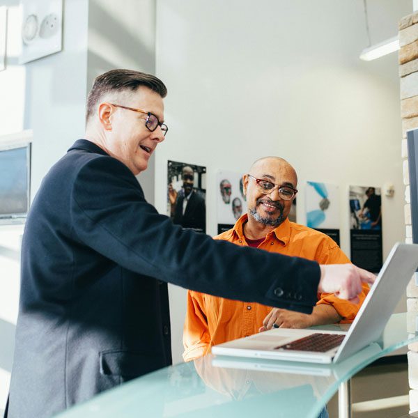 Man in glasses points to a laptop screen while another man in glasses looks on
