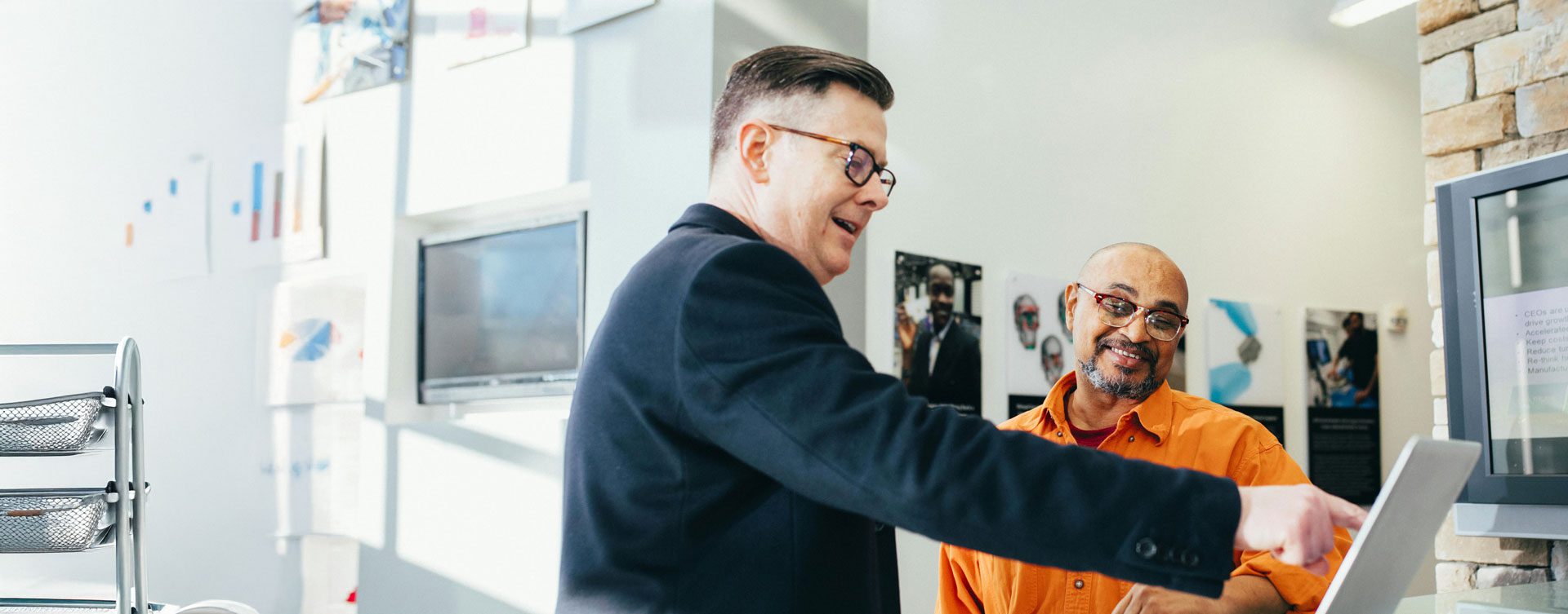 Two men stand in an office; one in a suit points at a laptop screen while the other, in an orange shirt, looks on and smiles.