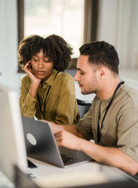 Two people sit at a desk looking at a laptop screen. One person points at the screen while the other rests her head on her hand and listens attentively.
