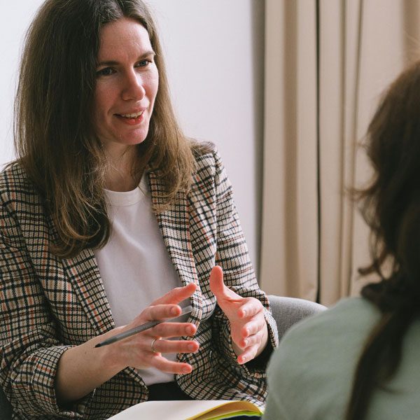 A dark haired woman holds a pen and explains something to a woman obscured in the foreground