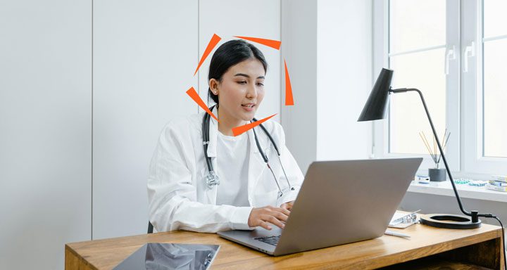 A doctor wearing a white coat and stethoscope sits at a desk, typing on a laptop in a well-lit office.