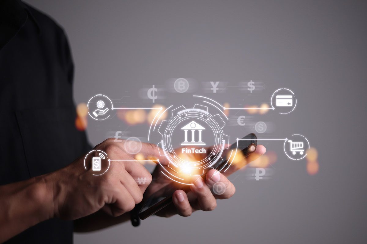 A person uses a smartphone with virtual icons representing financial technology, including currency symbols, bank, shopping cart, and security.