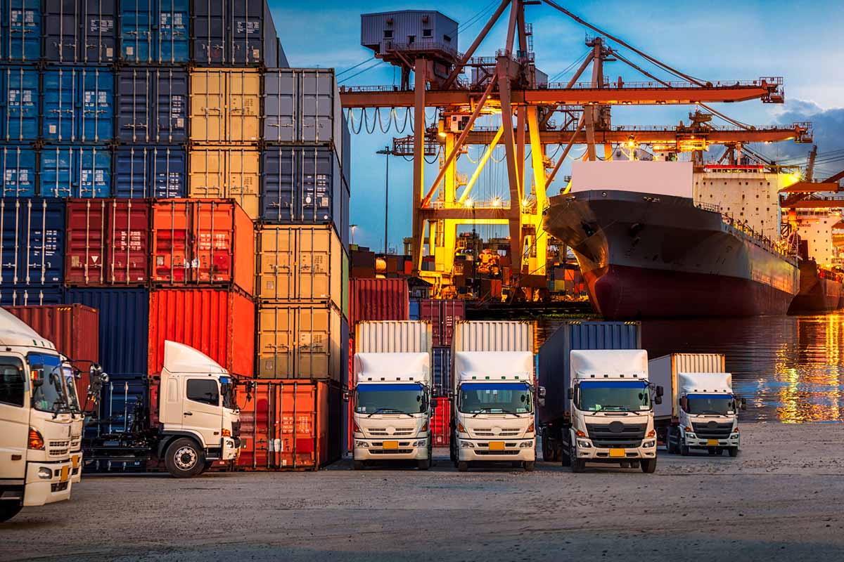 Rows of shipping containers, trucks, and cranes at a busy port, with a large cargo ship docked in the background during twilight.