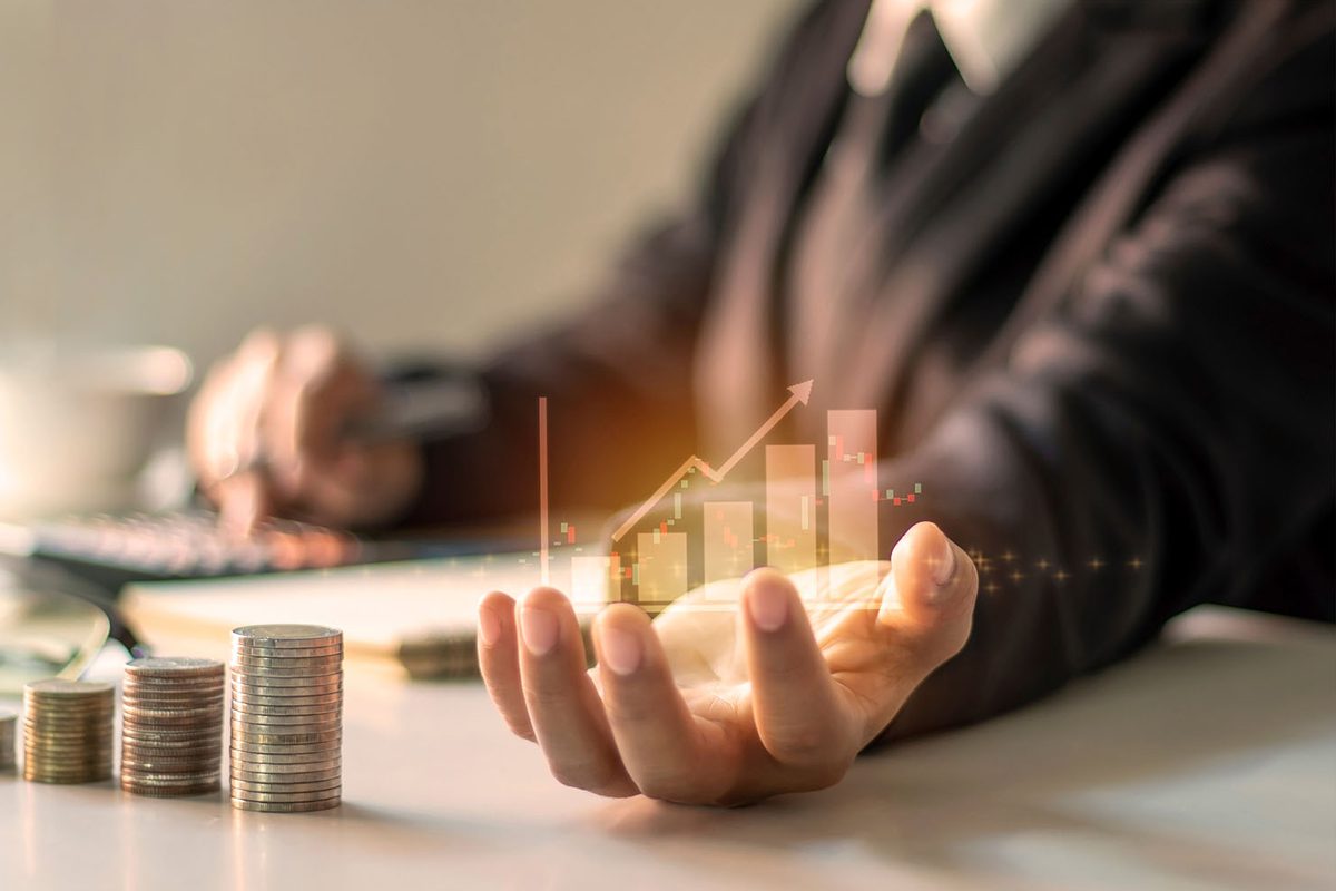 A person in a suit holds out a hand with a digital bar graph and arrow projection, next to stacked coins and a calculator on a desk.
