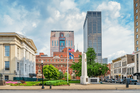 A downtown cityscape featuring historic and modern buildings, a flagpole with American flags, and a white monument in a landscaped area under a partly cloudy sky.