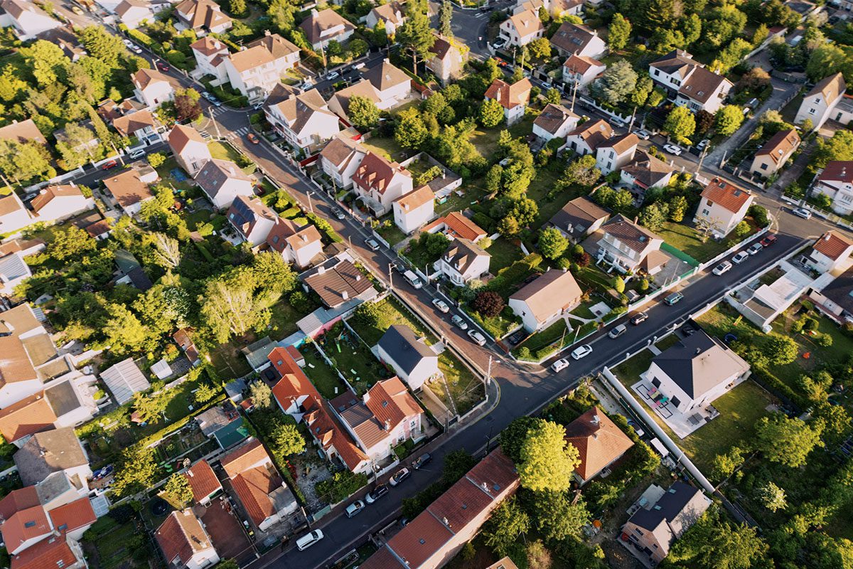Aerial view of a suburban neighborhood with tree-lined streets, detached houses, and parked cars along the roads.