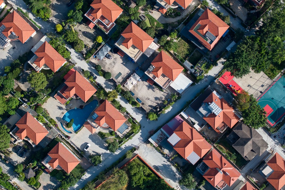 Aerial view of a suburban neighborhood with red-roofed houses, tree-lined streets, and a basketball court visible in the upper right corner.