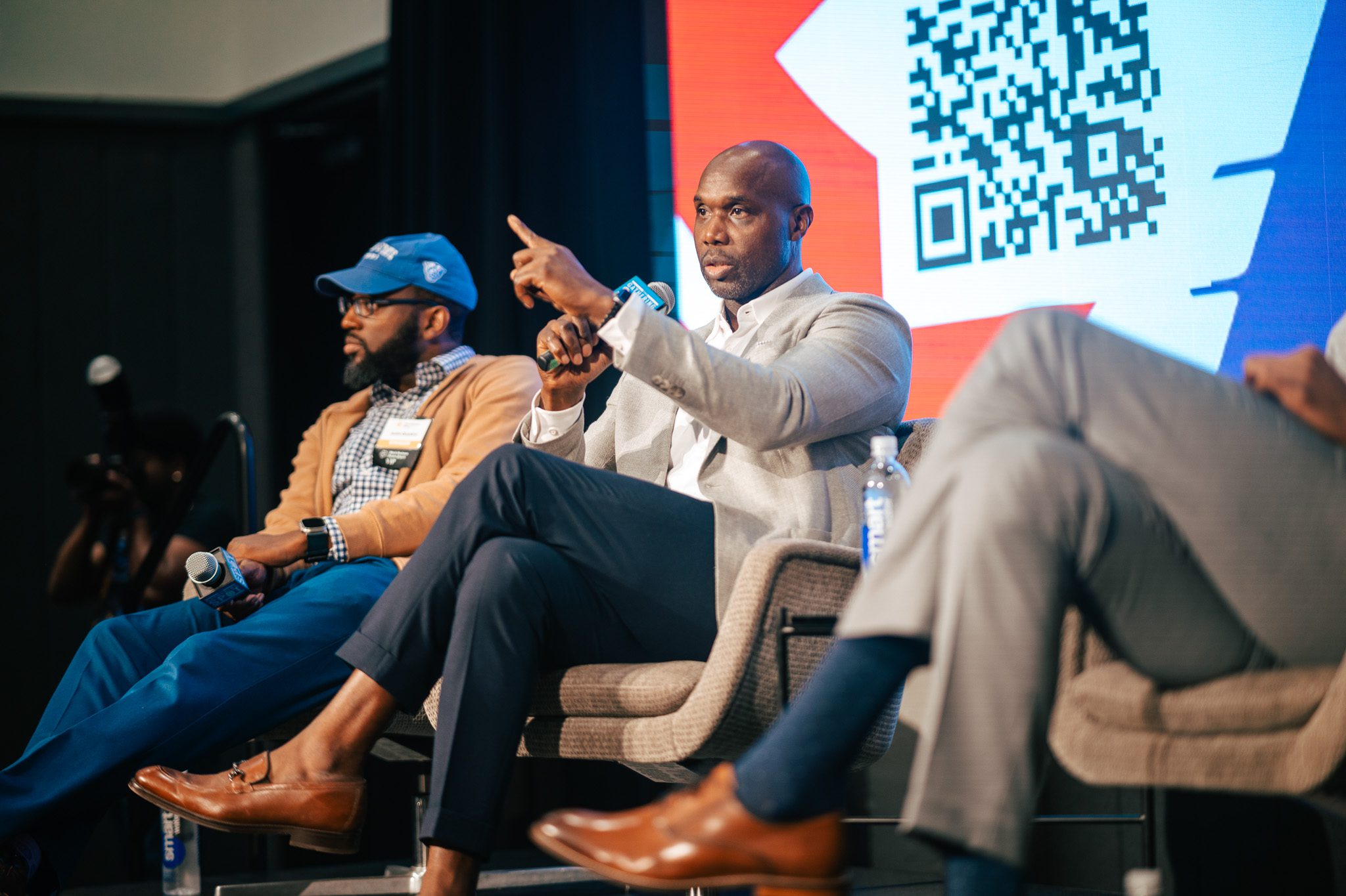 Three men are seated on a stage in discussion, with one man speaking into a microphone and gesturing; a QR code is displayed on a screen in the background.