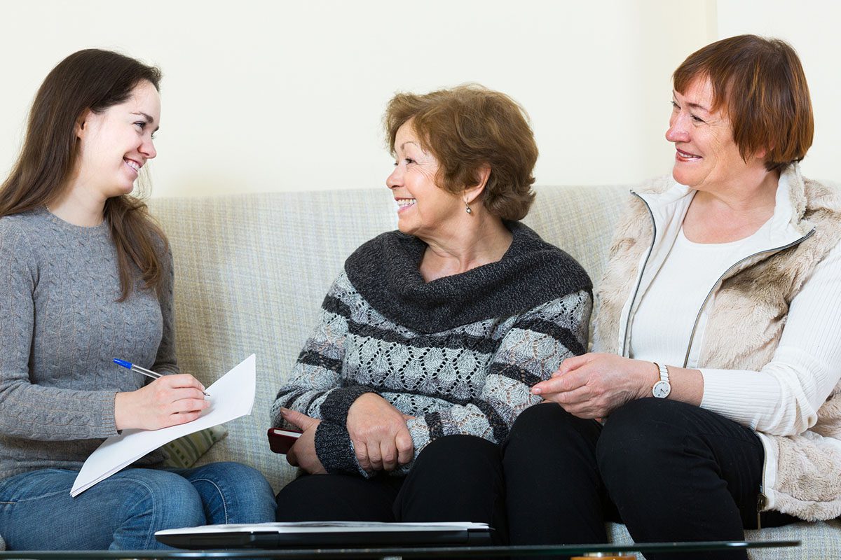 Three women sitting on a couch, two older and one younger, having a conversation. The younger woman holds a notepad and pen, while the others smile and listen.