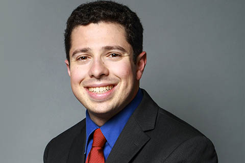 A man with short dark hair wearing a black suit, blue shirt, and red tie, smiling at the camera against a plain gray background.